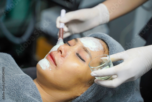 Closeup cosmetologist is applying facial white mask cream on woman client face in beauty clinic.