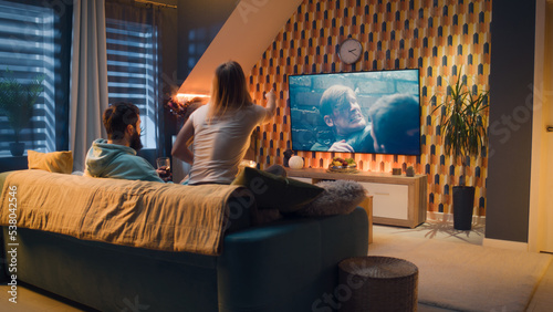 Bild auf Leinwand Happy couple sitting on sofa, watching action movie on TV or criminal blockbuster on streaming service, talking and discussing acting, eating snacks, pizza, chips