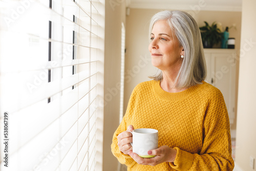 Thoughtful caucasian senior woman standing looking out of window at home with coffee, smiling