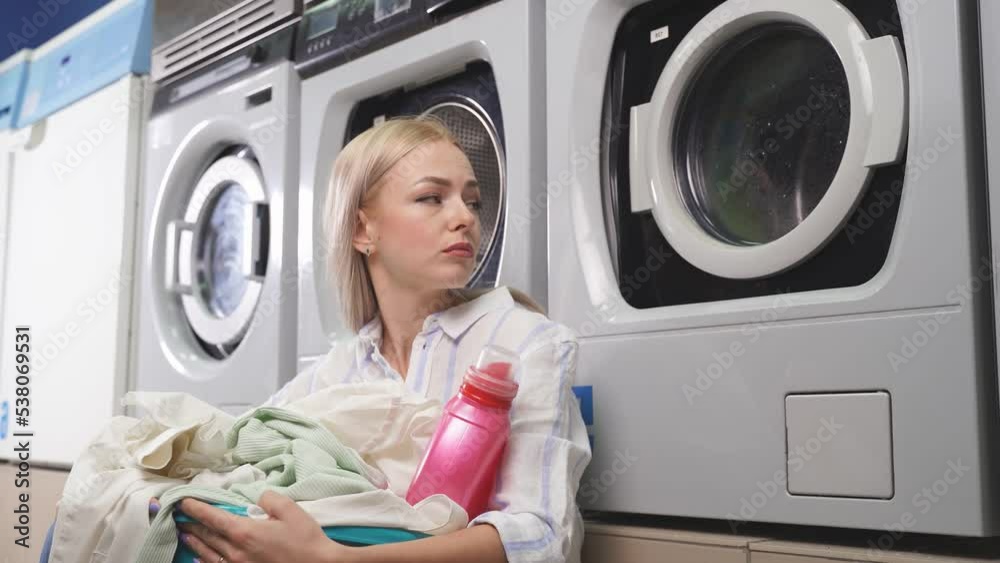 A woman is sitting near a household washing machine, which is located in a public laundry, she is waiting for clean laundry A woman is sitting near a household washing machine, which is located in a public laundry, she is waiting for clean laundry