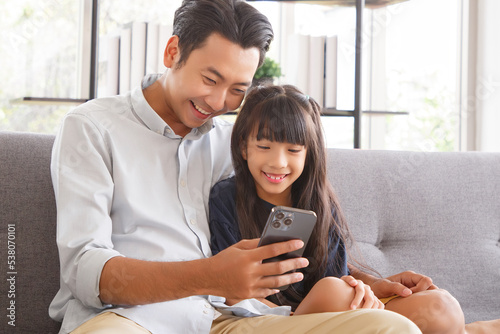 Cute asian girl help young father show something on smartphone making selfie together, smart daughter and dad sit on couch hold mobile phone take family picture on moblie phone.