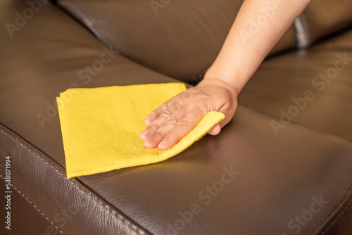 Shot of a woman cleaning black leather sofa with yellow rag at home