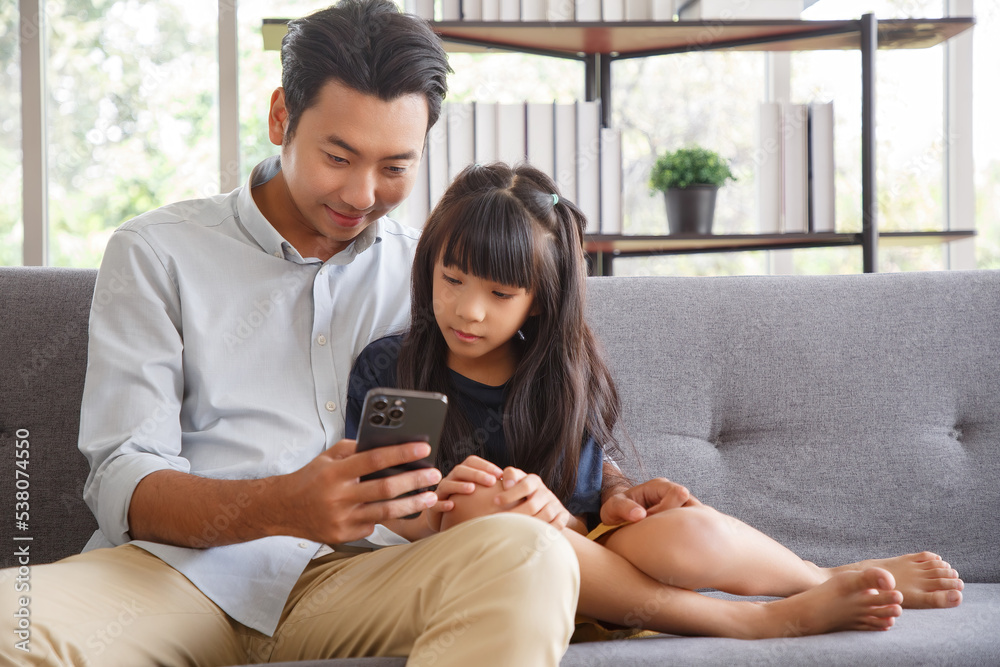 Cute asian girl help young father show something on smartphone making selfie together, smart daughter and dad sit on couch hold mobile phone take family picture on moblie phone. Cute asian girl help young father show something on smartphone making selfie together, smart daughter and dad sit on couch hold mobile phone take family picture on moblie phone.