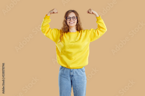 Cheerful teenage girl shows her biceps in funny way as sign of her strength, success and health. Portrait of young Caucasian woman in casual sweatshirt, jeans and glasses on beige background.