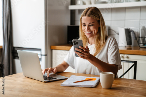 Happy woman using laptop and cellphone while sitting by table at home