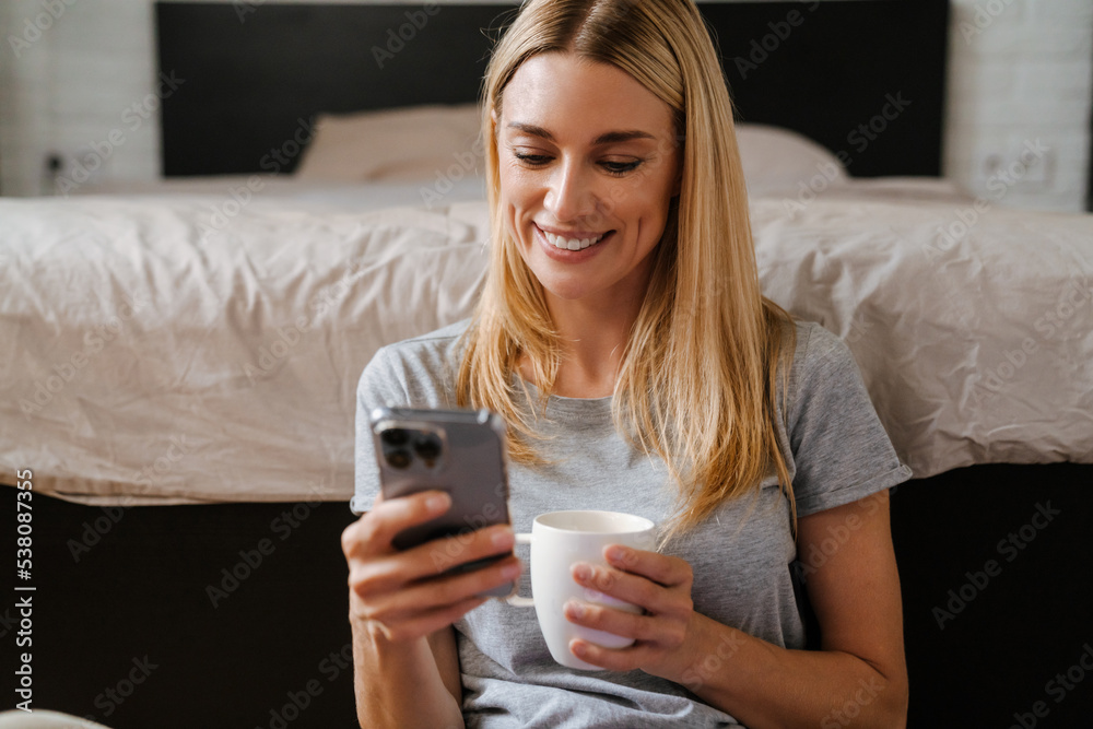 Blond woman drinking tea and using cellphone sitting over bed at home Blond woman drinking tea and using cellphone sitting over bed at home