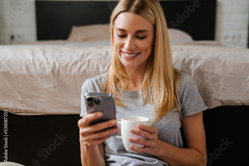 Blond woman drinking tea and using cellphone sitting over bed at home