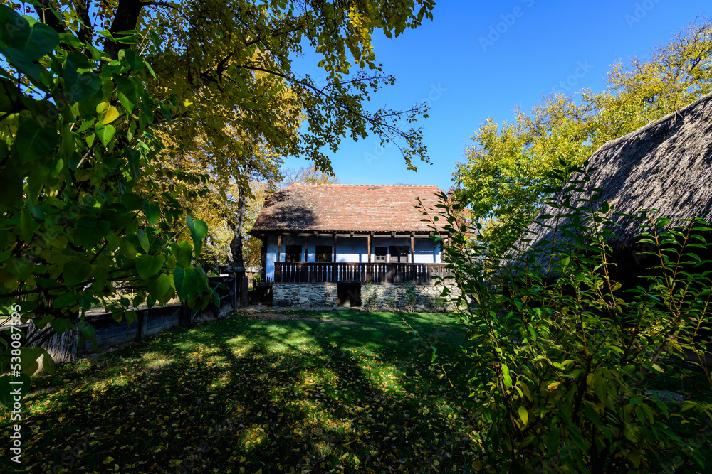 Traditional Romanian house surounded with many old trees with green, yellow, orange and brown leaves in Village Museum in Herastrau Park in Bucharest, Romania in a sunny autumn day. Traditional Romanian house surounded with many old trees with green, yellow, orange and brown leaves in Village Museum in Herastrau Park in Bucharest, Romania in a sunny autumn day.