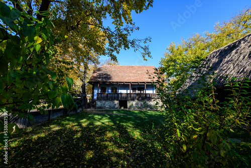 Traditional Romanian house surounded with many old trees with green, yellow, orange and brown leaves in Village Museum in Herastrau Park in Bucharest, Romania in a sunny autumn day.