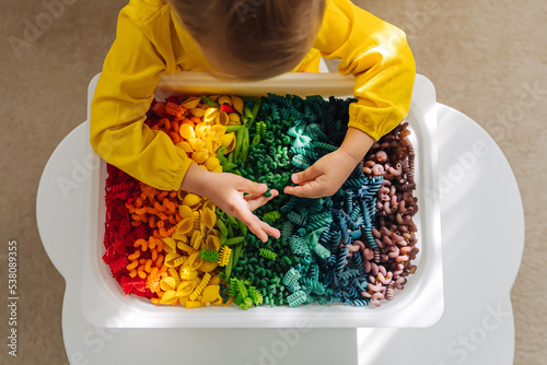 Child playing with sensory bin with dried pasta in rainbow colors. Dyed pasta for play and craft activities. Montessori material. Sensory play and learning colors activity for kids. photo