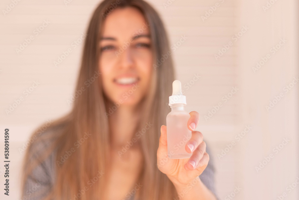 Selective focus of white and clear hair serum bottle in hand with blurred young woman in the background Selective focus of white and clear hair serum bottle in hand with blurred young woman in the background