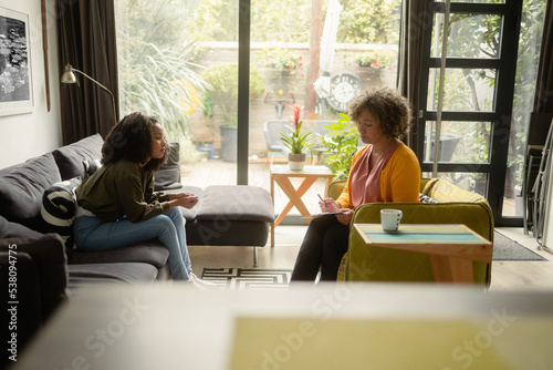 A teen African-American girl sits on a couch and talks to the female psychotherapist at her office. The girl looks like she's uncomfortable.