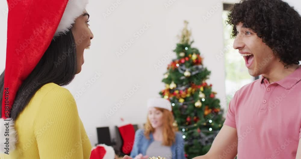 Happy biracial man giving present and hugging non binary transgender friend in santa hat at home Happy biracial man giving present and hugging non binary transgender friend in santa hat at home