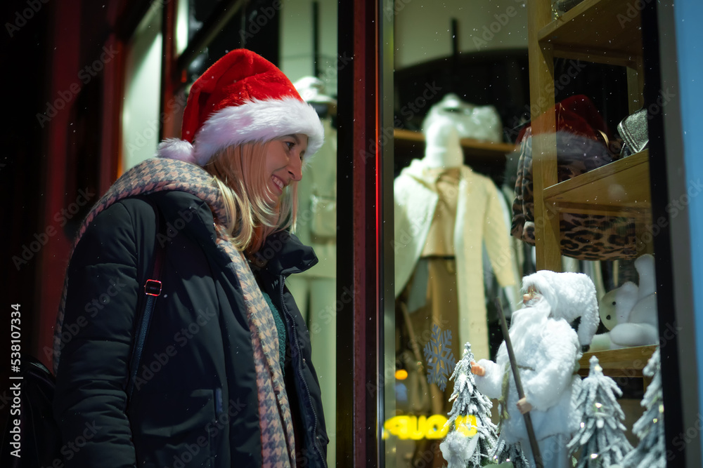 Young woman in a santa claus hat stands near the window, looks and smiles at a gift Santa Claus in a white suit on the street at night. Concept of holidays, gifts, happiness