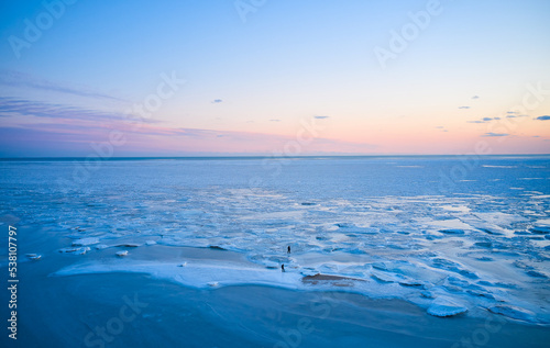 Aerial view - two people walk on ice on sunset over the frozen sea. Winter landscape on seashore during dusk. View from above of melting ice in ocean on sunrise. Global warming. Vivid colorful skyline