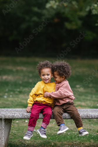 Black and Asian twins hugging on bench in park while smiling