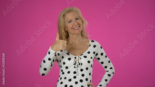 Promoter elderly blonde woman lady 50s in polka dot shirt gives thumbs up workspace copy space mockup promo area isolated on solid pink background studio portrait