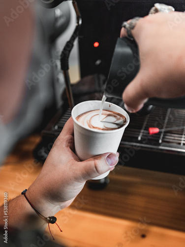 Barista pouring milk on white cup making a heart art on top of the coffe. Delicious espresso coffe preparation from a coffe shop. Hot organic drinks done. Espresso machine behind.