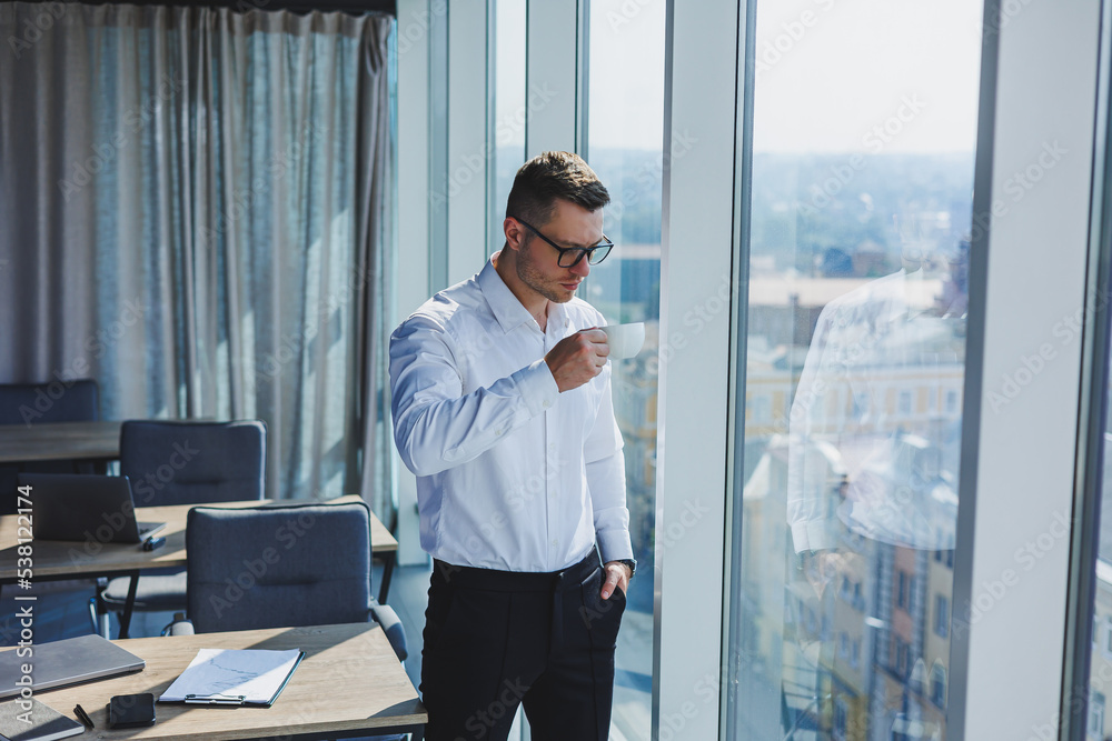 Fototapeta premium Cute young businessman with coffee in hand in glasses and white shirt, he is in modern office with big windows. A man in classic trousers and a white shirt. Young attractive businessman