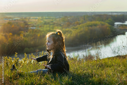 a little girl with curly hair sits on the ground on a walk outside the city in the rays of the setting sun photo