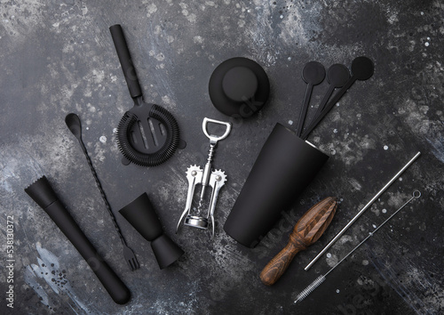 Black matt steel cocktail shaker,strainer,jigger and plastic stirrers with muddler on black stone background with manual wooden juicer.