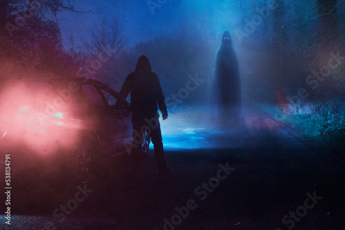 A supernatural concept of a spooky ghost floating above a road. With a driver next to car in a scary foggy winters forest at night photo