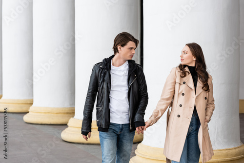 young man and woman in trench coat holding hands while walking near white columns.