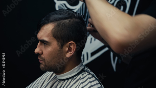 Man in hairdressing cape sitting near hairstylist in barbershop.
