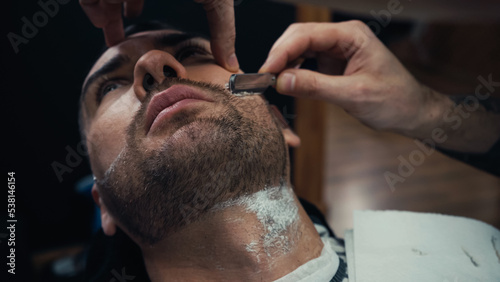 Barber shaving beard of client with razor in salon.