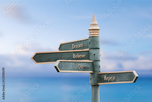 dream believe do repeat four word quote written on fancy steel signpost outdoors by the sea. Soft Blue ocean bokeh background.