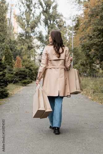 full length of young woman in trench coat holding shopping bags and walking in autumnal park.