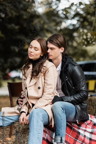 stylish man in black jacket hugging young girlfriend with closed eyes sitting in park.