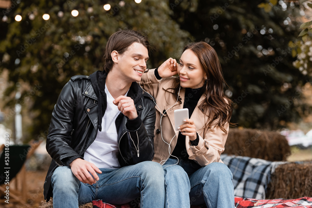 cheerful young woman holding smartphone while listening music and sharing wired earphones with boyfriend.