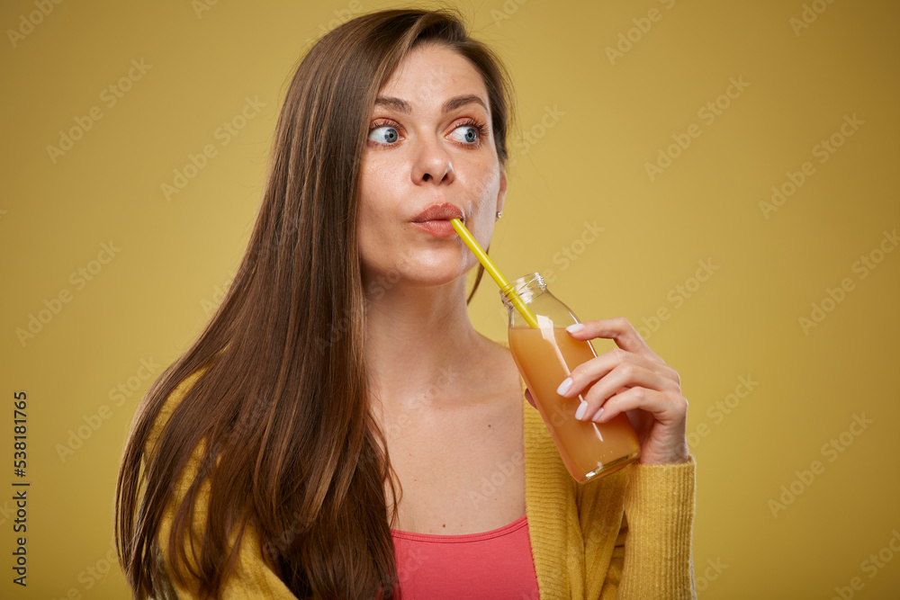 Fototapeta premium Young woman drinking orange juice from bottle with straw looking up. Close up face. Isolated advertising portrait on yellow.