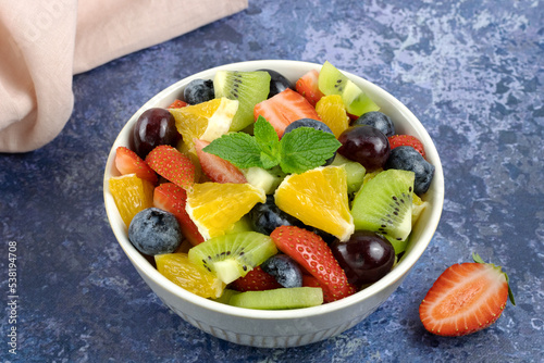 A bowl of juicy fruit and berry salad on a blue background. Fruit salad of strawberries, orange, kiwi, blueberries and grapes in a white bowl.