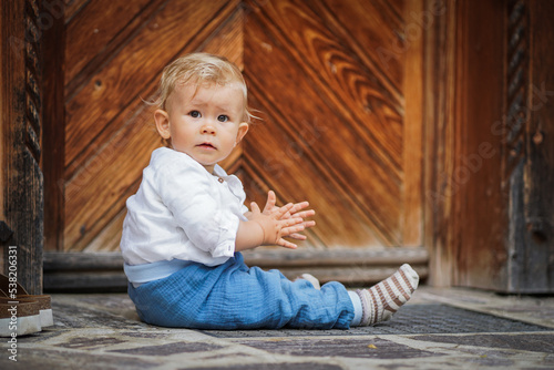 blond one year old baby boy in chic white shirt looking serious & focused into the camera outside of a rustic wooden hut while clapping hands and making hand signs