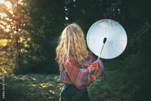 Beautiful shamanic girl playing on shaman frame drum in the nature.Shot from the back.