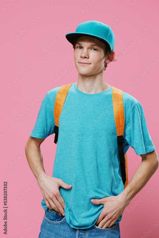 a handsome slender student guy is standing in a blue T-shirt and a cap with an orange backpack put on his shoulders on a pink background, putting his hands in his jeans pockets a handsome slender student guy is standing in a blue T-shirt and a cap with an orange backpack put on his shoulders on a pink background, putting his hands in his jeans pockets
