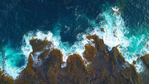 Footage of turquoise waves crashing over black and golden volcanic rocks on the coast