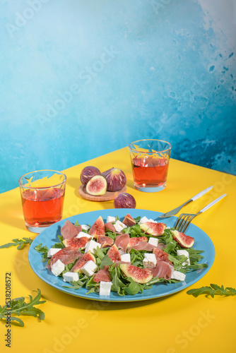 Autumn figs salad with arugula, feta, raw ham in blue plate on yellow and blue background served with rose wine. Flexitarian diet, paleo diet fall salad, copy space