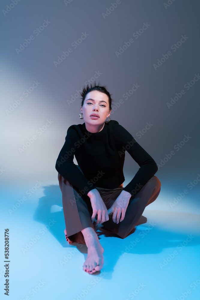 Obraz premium full length of stylish barefoot woman in black sweater looking at camera while sitting on grey and blue background.