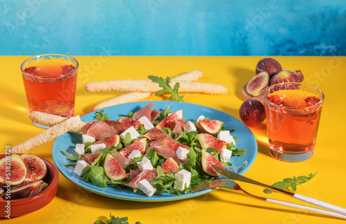 Autumn figs salad with arugula, feta, raw ham in blue plate on yellow and blue background served with rose wine. Flexitarian diet, paleo diet fall salad, close up