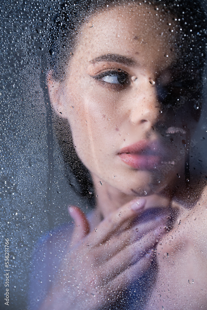 Young brunette woman touching neck behind wet glass on grey background.