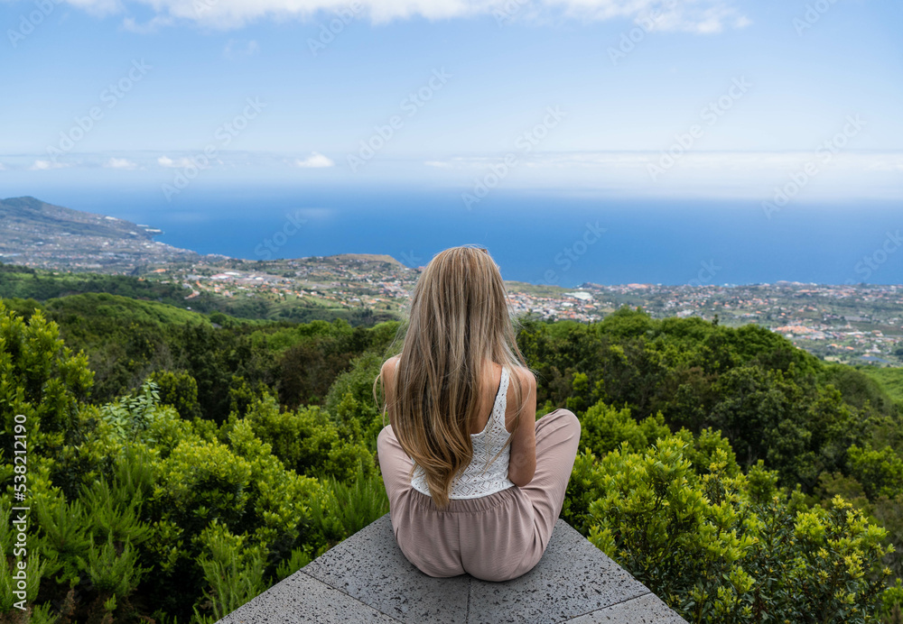 Backwards of caucasian woman with long blonde hair at the viewpoint of the town in the middle of the nature