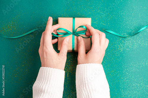 Human hands in white sweater tieding brown gift box with a emerald green satin ribbon bow on green background with gold colored glitter Christmas Holidays concept flat lay