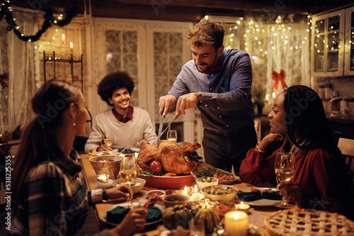 Happy man carving Thanksgiving turkey during dinner party with friends.