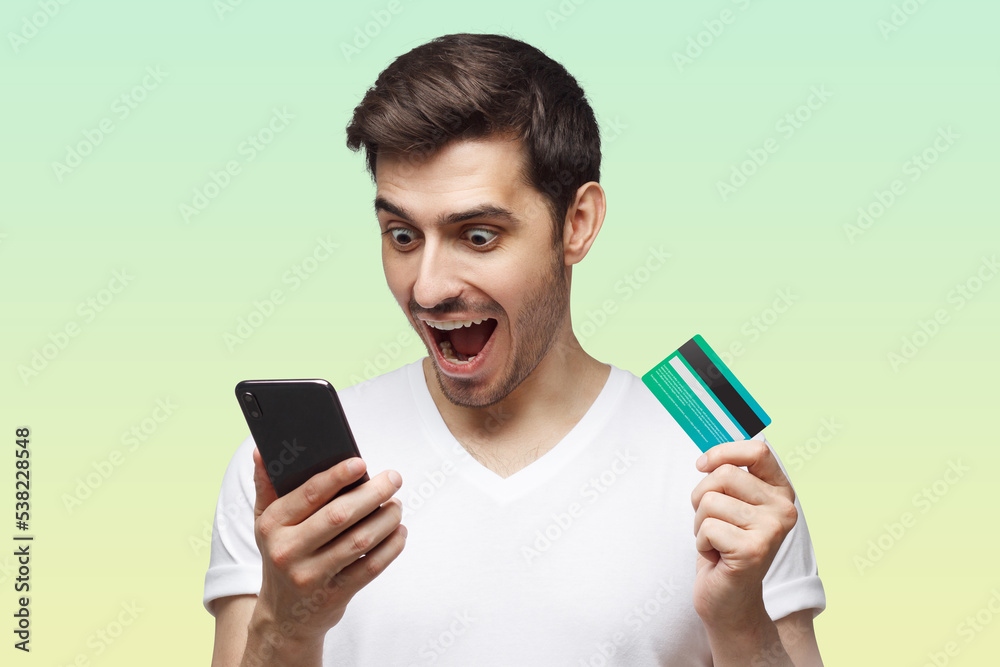 Excited handsome man with mouth open, holding credit card, using mobile phone on green background Excited handsome man with mouth open, holding credit card, using mobile phone on green background
