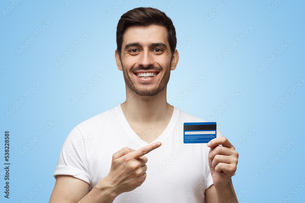 Handsome smiling young man in white t-shirt showing blue credit card, pointing on it with finger Handsome smiling young man in white t-shirt showing blue credit card, pointing on it with finger