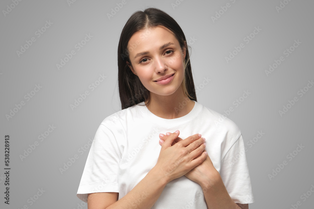Smiling positive woman keeps hands on chest, expresses sympathy wearing blank white t-shirt Smiling positive woman keeps hands on chest, expresses sympathy wearing blank white t-shirt