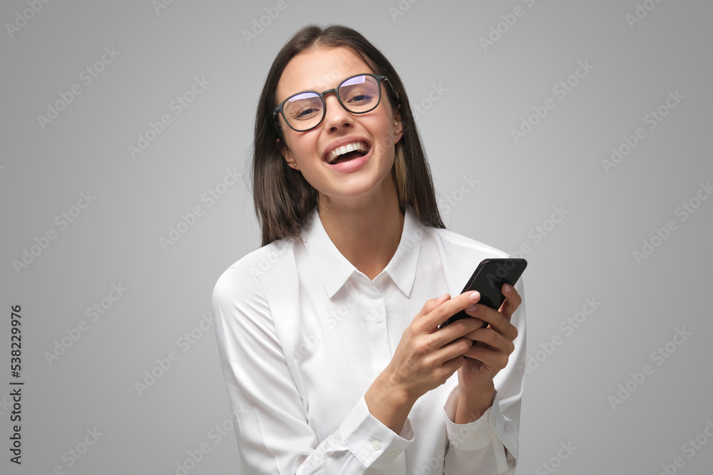 Positive laughing lady in glasses and white T-shirt holding smartphone, isolated on gray Positive laughing lady in glasses and white T-shirt holding smartphone, isolated on gray